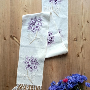 May include: A long, off-white fabric table runner featuring embroidered purple floral motifs with intricate stitching. The runner has a decorative knotted fringe at the bottom edge and is displayed vertically, showcasing multiple repeating flower patterns.