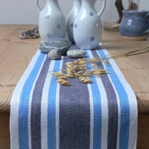 May include: A table runner featuring vertical stripes in shades of light blue, dark blue, and white. The runner is decorated with three white ceramic pitchers with blue floral patterns, several smooth grey stones, and a sprig of dried oat stalks.