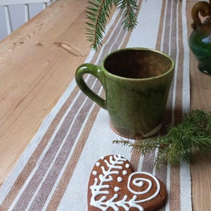 May include: A green ceramic mug with a handle rests on a striped table runner alongside a heart-shaped gingerbread biscuit decorated with white icing. A sprig of evergreen adds a festive touch, suggesting a holiday theme.