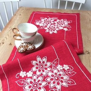 Könnte beinhalten: Roter, bestickter Tischläufer mit weißem Blumendesign. Zwei Teetassen mit goldenem Rand und ein Teller mit Keksen stehen auf dem Tisch. Der Läufer ist rechteckig und hat einen weißen Rand.