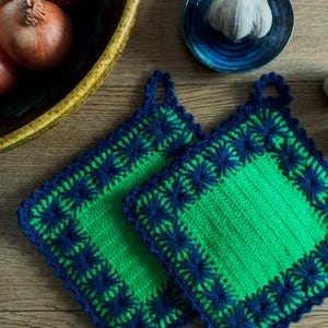 Può includere: Due presine quadrate fatte a mano all'uncinetto. Le presine sono verdi con un bordo blu scuro e un motivo a stella. Sono su una superficie di legno, con una ciotola di cipolle e un piatto blu con aglio sullo sfondo.