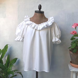 May include: White cotton blouse with a large ruffle collar and puffed sleeves. The blouse is on a mannequin and has a relaxed fit.