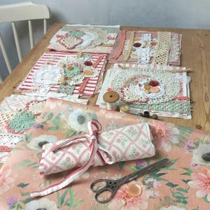 Op de afbeelding: Een verzameling handgemaakte textielkunstwerken, waaronder doilies, stofresten en versieringen zoals knopen en kant, gerangschikt op een houten tafel. Een stof met bloemenpatroon en een schaar zijn ook aanwezig.