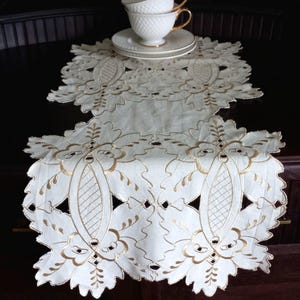 May include: An off-white table runner featuring intricate cutwork and gold-toned embroidery in a floral and leaf pattern. Two white teacups with gold rims and a textured surface are stacked on a matching saucer atop the runner.