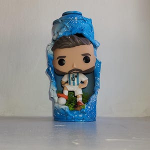 Peut inclure: Figurine Funko Pop d'un joueur de football en uniforme blanc et bleu, numéro 10, debout sur un morceau d'herbe verte. La figurine est enfermée dans une structure bleue semblable à de l'eau avec des accents blancs.