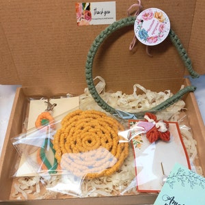May include: A gift box filled with handmade macrame items, including a headband, a wall hanging, and a small decorative piece. The box is lined with brown paper and the items are wrapped in clear plastic. The box also includes a small card with the text "Amaranth macrame Made by Teddi Stankova Handmade with love".