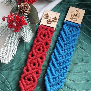 Op de afbeelding: Twee macramé armbanden, één rood en één blauw, met houten labels met kerstthema's. De rode armband heeft een cirkelpatroon, de blauwe een visgraatpatroon. Feestelijke vakantiedecoratie op de achtergrond.
