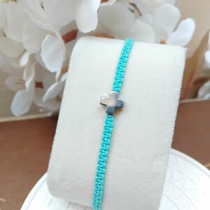 May include: A turquoise macrame bracelet featuring a silver cross charm. The bracelet is presented on a white, rectangular surface, with white flowers in the background. The cross is positioned centrally.