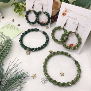 Könnte beinhalten: Drei handgefertigte Schmuckstücke aus grünem Garn mit goldenen Akzenten. Die Stücke umfassen ein Paar Ohrringe, ein Armband mit einem Sternanhänger und ein Armband mit einem Schneeflockenanhänger. Der Schmuck wird auf weißen Karten mit dem aufgedruckten Text "Amaranth Madame" präsentiert.