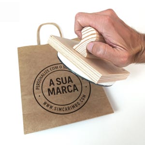 Puede incluir: Un sello de madera con mango se presiona sobre una bolsa de papel marrón. El sello tiene un diseño circular con el texto "A SUA MARCA" y "WWW.SIMCARIMBO.COM". La bolsa tiene un asa y está sobre un fondo blanco.