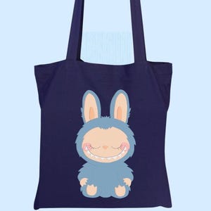 May include: A navy blue tote bag with a cartoon monster-bunny design. The character is light blue with a smiling face and large ears. The bag has two navy blue handles and is set against a light blue background.