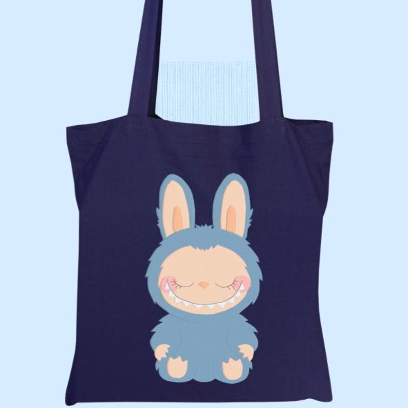 May include: A navy blue tote bag with a cartoon monster-bunny design. The character is light blue with a smiling face and large ears. The bag has two navy blue handles and is set against a light blue background.