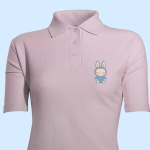 May include: A pale purple polo shirt with short sleeves and a collared neckline. The shirt has a small, blue cartoon bunny graphic on the left chest. It has three buttons at the placket.