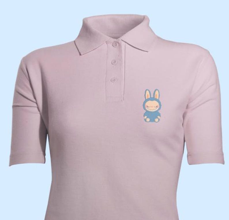 May include: A pale purple polo shirt with short sleeves and a collared neckline. The shirt has a small, blue cartoon bunny graphic on the left chest. It has three buttons at the placket.