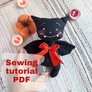 Op de afbeelding: Naaihandleiding PDF voor het maken van een zwart vleermuis knuffel met rood lint. De vleermuis heeft een gestikt gezicht met witte ogen en tanden.