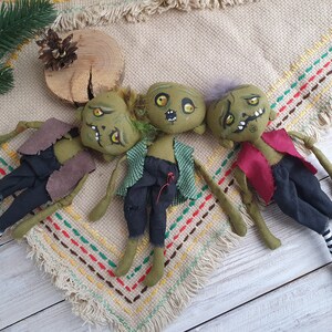 K&ouml;nnte beinhalten: Drei gr&uuml;ne Stoffpuppen mit gestickten Gesichtern und bemalten Details. Die Puppen sind in verschiedenen Outfits gekleidet, darunter eine braune Weste, ein kariertes Hemd und ein rotes Hemd. Die Puppen liegen auf einem beige-braunen gemusterten Stoff.