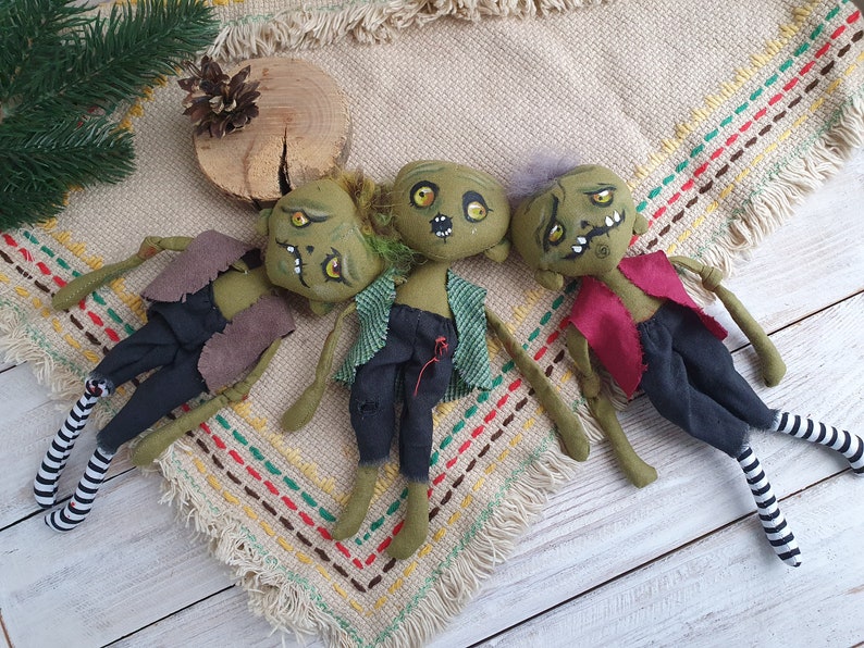 K&ouml;nnte beinhalten: Drei gr&uuml;ne Stoffpuppen mit gestickten Gesichtern und bemalten Details. Die Puppen sind in verschiedenen Outfits gekleidet, darunter eine braune Weste, ein kariertes Hemd und ein rotes Hemd. Die Puppen liegen auf einem beige-braunen gemusterten Stoff.