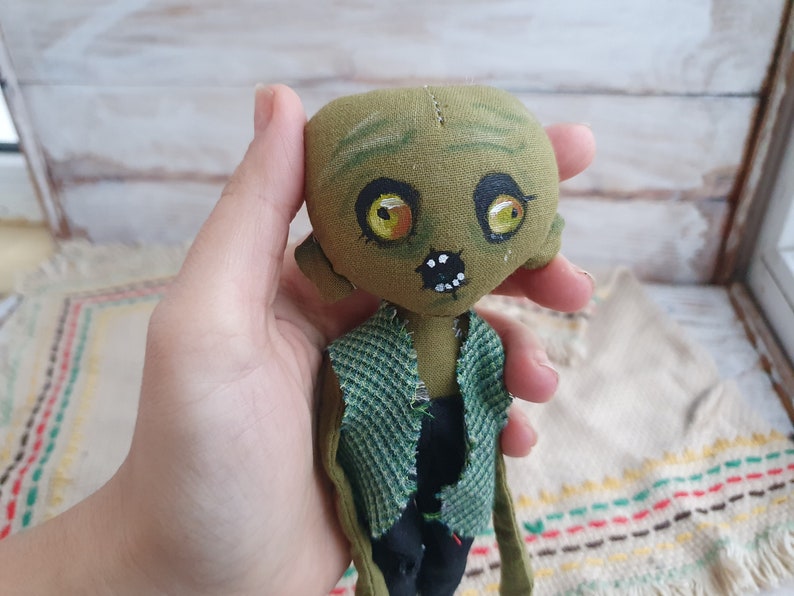 K&ouml;nnte beinhalten: Eine gr&uuml;ne Stoffpuppe mit bemalten Gesichtsz&uuml;gen, darunter gelbe Augen, ein schwarzer Mund und wei&szlig;e Z&auml;hne. Die Puppe tr&auml;gt ein gr&uuml;n-schwarzes kariertes Hemd.