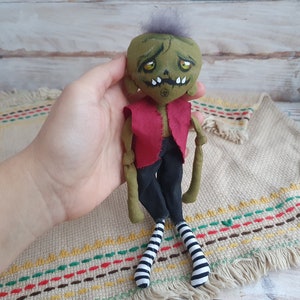 K&ouml;nnte beinhalten: Eine gr&uuml;ne und schwarze Stoffpuppe mit einem roten Shirt und schwarz-wei&szlig;en gestreiften Socken. Die Puppe hat ein bemaltes Gesicht mit gr&uuml;ner Haut, gelben Augen und einem zahnigen Grinsen.