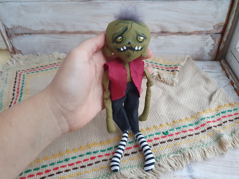 K&ouml;nnte beinhalten: Eine gr&uuml;ne und schwarze Stoffpuppe mit einem roten Shirt und schwarz-wei&szlig;en gestreiften Socken. Die Puppe hat ein bemaltes Gesicht mit gr&uuml;ner Haut, gelben Augen und einem zahnigen Grinsen.