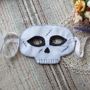 May include: White felt skull mask with black felt eyes and stitched details. The mask has white ribbon ties.