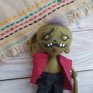 K&ouml;nnte beinhalten: Eine gr&uuml;ne Stoff-Zombie-Puppe mit einem roten Shirt, einer schwarzen Hose und einem bemalten Gesicht. Die Puppe hat eine stachelige graue Frisur und einen zusammengen&auml;hten Mund.