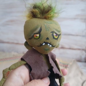 K&ouml;nnte beinhalten: Eine handgemachte gr&uuml;ne Zombiedoll mit gelben Augen und einem braunen, zerrissenen Hemd. Die Puppe hat einen gen&auml;hten Mund und eine unordentliche gr&uuml;ne Frisur.
