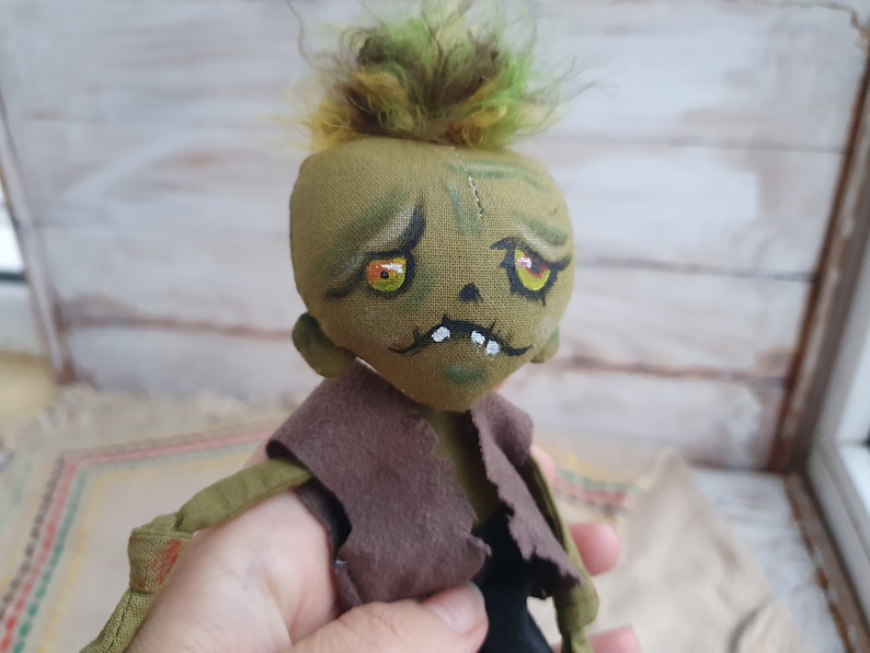 K&ouml;nnte beinhalten: Eine handgemachte gr&uuml;ne Zombiedoll mit gelben Augen und einem braunen, zerrissenen Hemd. Die Puppe hat einen gen&auml;hten Mund und eine unordentliche gr&uuml;ne Frisur.