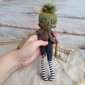 K&ouml;nnte beinhalten: Eine handgemachte gr&uuml;ne Zombie-Puppe mit schwarz-wei&szlig;en gestreiften Str&uuml;mpfen, brauner Weste und gr&uuml;nem Haar. Die Puppe hat ein bemaltes Gesicht mit gelben Augen und einem zahnigen Grinsen.