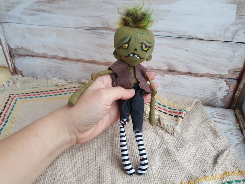 K&ouml;nnte beinhalten: Eine handgemachte gr&uuml;ne Zombie-Puppe mit schwarz-wei&szlig;en gestreiften Str&uuml;mpfen, brauner Weste und gr&uuml;nem Haar. Die Puppe hat ein bemaltes Gesicht mit gelben Augen und einem zahnigen Grinsen.