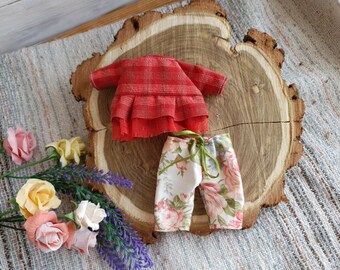 Puede incluir: Un vestido de cuadros rojos y blancos con un dobladillo con volantes y un pantalón de estampado floral. La ropa está colocada sobre una superficie de madera.