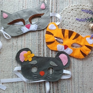 Peut inclure: Trois masques d'animaux en feutre avec des élastiques blancs. Les masques sont un chat gris, un tigre orange et une souris grise avec une oreille rose et une fleur jaune.