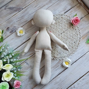 May include: A simple, unpainted doll made of beige fabric with a round head and long limbs. The doll is lying on a white wooden surface with a lace doily and artificial flowers.