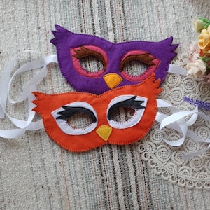 May include: Two felt owl masks, one orange and one purple, with white eye details and black eyelashes. The masks have white ribbon ties.