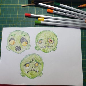 K&ouml;nnte beinhalten: Drei Cartoon-Zeichnungen von gr&uuml;nen Zombie-Gesichtern mit verschiedenen Ausdr&uuml;cken. Die Gesichter sind mit Bleistift auf wei&szlig;em Papier gezeichnet.