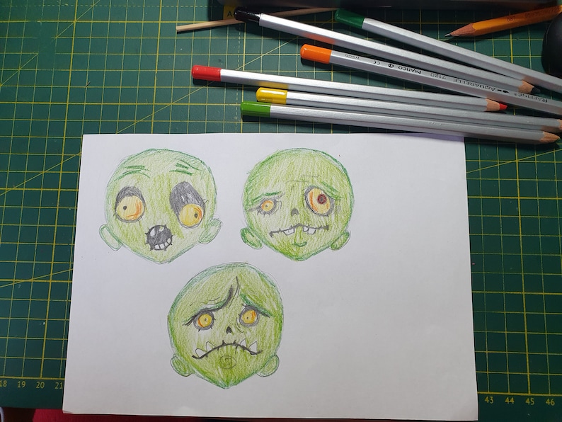 K&ouml;nnte beinhalten: Drei Cartoon-Zeichnungen von gr&uuml;nen Zombie-Gesichtern mit verschiedenen Ausdr&uuml;cken. Die Gesichter sind mit Bleistift auf wei&szlig;em Papier gezeichnet.