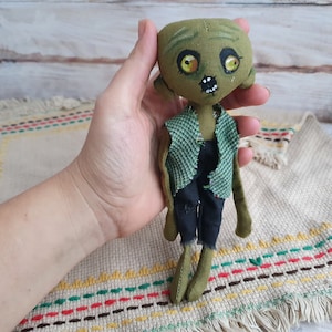 K&ouml;nnte beinhalten: Eine gr&uuml;ne Stoffpuppe mit einem abgenutzten Look, die ein gr&uuml;nes und schwarzes Outfit tr&auml;gt. Die Puppe hat bemalte Augen und einen gestickten Mund.