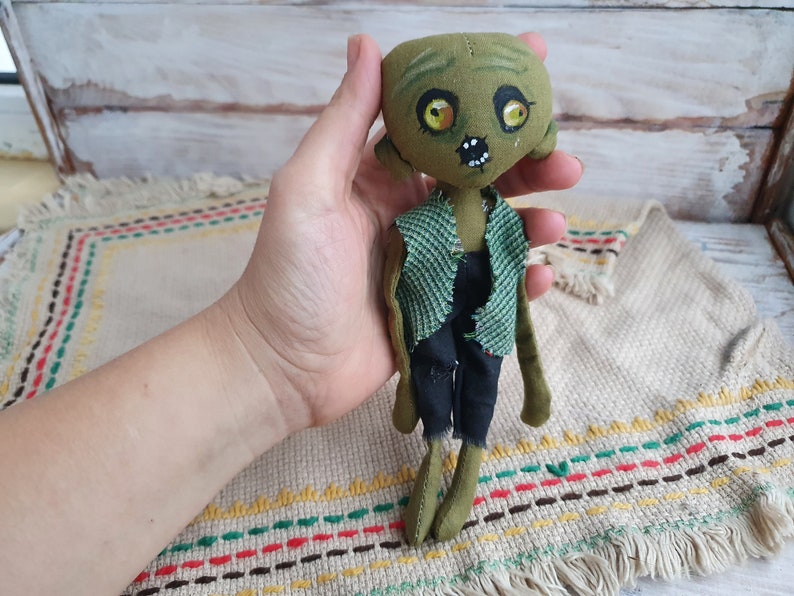 K&ouml;nnte beinhalten: Eine gr&uuml;ne Stoffpuppe mit einem abgenutzten Look, die ein gr&uuml;nes und schwarzes Outfit tr&auml;gt. Die Puppe hat bemalte Augen und einen gestickten Mund.