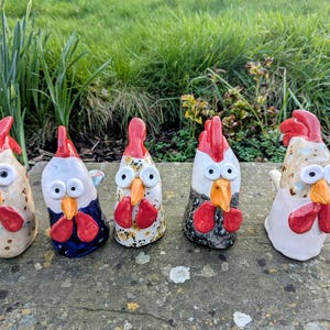 May include: Five ceramic rooster ornaments with red crests, orange beaks, and large eyes. The roosters feature diverse colour schemes, including white, blue, and speckled designs. They are displayed on a stone surface with a green backdrop.