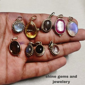 以下が含まれることがあります： ムーンストーン、シトリン、ルビーなど、さまざまな形と色の宝石ペンダントのコレクション。各ペンダントには、チェーンに取り付けるためのシルバー色のベイルが付いています。画像には「shine gems and jewelery」というテキストが含まれています。