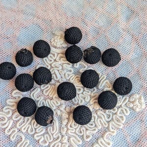 May include: A selection of small, round, black fabric-covered buttons. The buttons are displayed on a patterned, off-white and pastel-coloured lace fabric. The buttons have a textured, woven appearance.