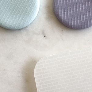 May include: Three textured, round, silicone mats in light blue, purple and white. The mats have a brick pattern.
