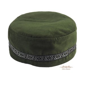 May include: A round, olive green corduroy hat with a flat top and a structured side band. The side of the hat features a black trim decorated with a repeating white geometric pattern. The fabric has a visible ribbed texture throughout the surface.