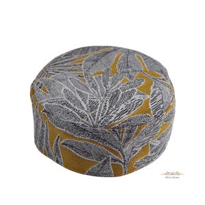 May include: A round, brimless cap featuring a woven fabric with a mustard yellow background and a dense, repeating pattern of dark gray and white stylized leaves. The structure is cylindrical with a flat top, displaying a textured textile finish.