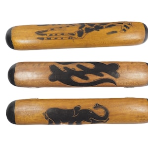 Puede incluir: Tres instrumentos de percusión cilíndricos de madera con extremos redondeados negros. El instrumento superior presenta una ilustración de jirafa, el central muestra un símbolo abstracto negro y el inferior un gráfico de elefante, todo en tinta negra.