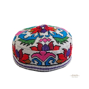 Puede incluir: Un gorro redondo de parte superior plana con un intrincado bordado de punto de cruz sobre un fondo gris claro. El diseño muestra vibrantes patrones florales y geométricos en tonos rojo, azul, verde, morado y rosa, con un borde morado oscuro.