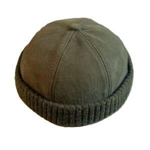May include: An olive green docker cap featuring a smooth, suede-like crown with six stitched panels meeting at the centre. The brim consists of a thick, ribbed knit band that encircles the base, providing a textured contrast to the crown.