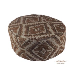 Puede incluir: Un gorro redondo de parte superior plana con un patrón geométrico de diamantes en color marrón y crema tejido en la tela. El diseño consiste en formas angulares repetitivas en toda la superficie de la prenda. El texto Ethno-Bazaar es visible.