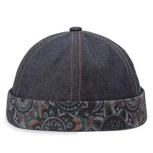 May include: A dark blue denim docker cap featuring a rolled brim with a multi-colored paisley pattern in shades of green, orange, and white. The crown is constructed from six panels of dark blue denim fabric with visible copper-colored stitching.