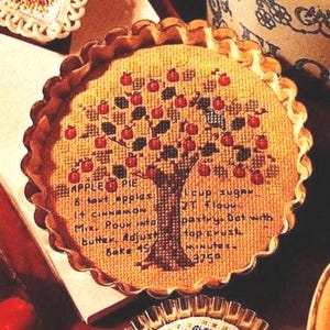 Può includere: Un ricamo a punto croce rotondo raffigurante un melo con frutti rossi e foglie verdi. Il testo recita: APPLE PIE, 6 tart apples, 1 cup sugar, 1 t cinnamon, 2 T flour, Mix. Pour into pastry. Dot with butter. Adjust top crust. Bake 45 minutes 375 degrees.